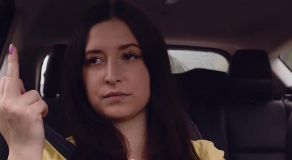 Peggy, wearing a yellow shirt, is sitting in the back seat of a car.